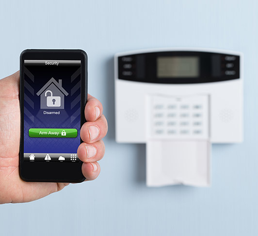 A person holding a smart phone next to a home security system.